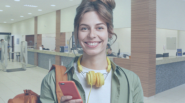 Foto ilustrativa de uma mulher em uma sala, ela está de pé e sorrindo com fone no pescoço e um celular nas mãos.