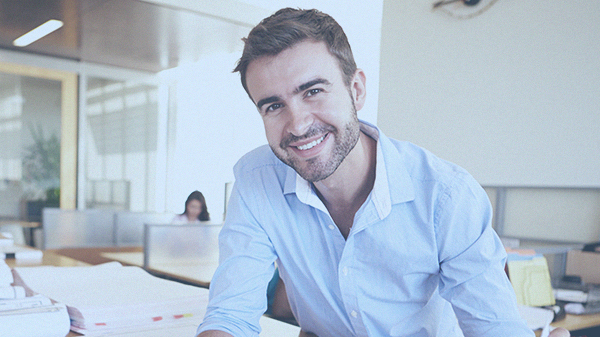 Foto ilustrativa de um homem sorrindo, camisa social clara em um grande escritório.